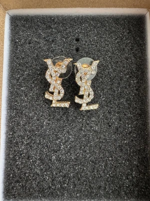 YSL örhängen med strass. - Snygga YSL örhängen i guld med glittrande strass-stenar. Ikoniska logotypen i en stilren design som verkligen sticker ut. Perfekta för dig som vill ha något extra till din look. Kommer med klassisk plupp-bakdel för enkel användning. Aldrig använda.