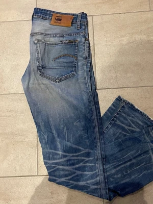 Blå jeans från G-Star RAW - Säljer ett par blå jeans från G-Star RAW med snyggt slitna detaljer och ljusa tvättade partier. Klassisk femficksmodell med läderpatch bak i midjan och raka ben. Jeansen har kontrastsömmar och coola whiskers-effekter på benen. Perfekta till sneakers och hoodie. 32/34