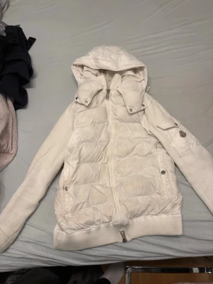 Vit Moncler pufferjacka med huva - Säljer en vit cardigan jacka från Moncler med avtagbar huva och ribbade stickade ärmar. Jackan har dragkedja framtill, fickor med tryckknappar och en ikonisk Moncler-logga på ärmen. Insidan har ett coolt serietidningsmönster. Perfekt för kalla dagar! Den har storlek L men passar mindre.