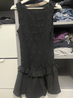 Svart kort klänning med volang - Säljer en svart kort klänning med smockad överdel och volangdetaljer vid kjolkanten. Klänningen är ärmlös och har en rund halsringning. Materialet känns lätt och luftigt, perfekt för varma dagar eller när du vill känna dig extra bekväm.