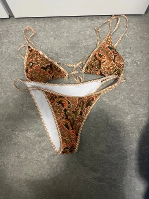 Paisleymönstrad bikini i brunt och orange - Säljer en snygg bikini med paisleymönster i brunt, orange och grönt. Överdelen har trekantskupor med smala axelband och knyt i ryggen. Underdelen är högt skuren med matchande mönster och smala sidor. Perfekt för strandhäng eller poolparty!