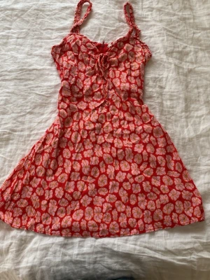 Röd rosa blommig kortklänning med knyt - Supersöt röd miniklänning med rosa blommönster och smala axelband. Klänningen har en fin knytning framtill vid bysten och dragkedja. 