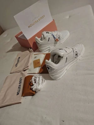 Vita Louis Vuitton sneakers med logga - Säljer ett par riktigt snygga Louis Vuitton sneakers i helvitt skinn med klassisk LV-logga på plösen och sidan. Skorna har vita snören, platt sula och diskreta svarta detaljer. Kommer med originalkartong, extra skosnören och dustbags.