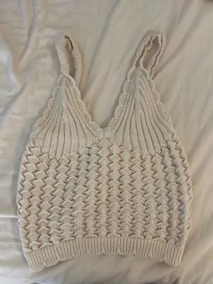 Beige stickad linnetopp - Säljer en beige stickad linnetopp med breda axelband och v-ringad hals. Toppen har ett snyggt virkat mönster och ribbade detaljer längs kanterna. Perfekt för sommarens alla tillfällen och superfin att styla med jeans eller kjol.