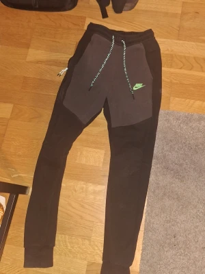 Svarta Nike mjukisbyxor med snörning - Säljer ett par svarta mjukisbyxor från Nike med kontrasterande mörkgrå paneler framtill och grönt Nike-logga på vänster sida. Har stor ficka på sidan, kan visa bild i chatten.