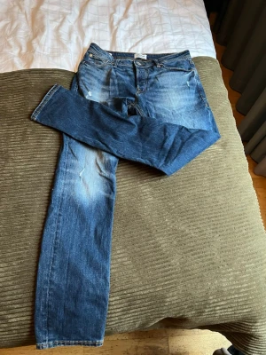 Blå jeans från Jack & Jones - Säljer ett par klassiska blå jeans från Jack & Jones med snygg slitning och ljusare partier på låren och knäna. Jeansen har rak passform, fem fickor och normal midja. Perfekta till en avslappnad stil och funkar året runt.
