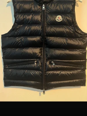 Svart dunväst från Moncler - Säljer en svart dunväst från Moncler Longue Saison. Västen har quiltad design, två dragkedjefickor fram och Moncler-logga på bröstet. Stängs med dragkedja och har en clean, sportig look. Perfekt för lager-på-lager under kyliga dagar.
