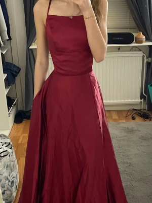 Vinröd balklänning med öppen rygg - Säljer en vinröd långklänning med snygg snörning i ryggen och öppen rygg. Den är aldrig använd och har prislapp. Två fickor med en slit vid benet. Perfekt för speciella tillfällen och bal💕