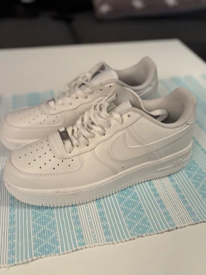 Nike Air Force 1 vita sneakers - Klassiska Nike Air Force 1 sneakers i helvitt skinn med perforerad tå, rund tå och vit sula. Skorna har snörning och den ikoniska Nike-loggan på sidan och hälen. Perfekta för dig som vill ha en clean och tidlös look.