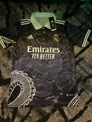Real Madrid fotbollströja - Säljer en svart Real Madrid t-shirt från Adidas med grymt drakmönster över hela framsidan. Tröjan har vit krage, korta ärmar och broderade loggor. Materialet är polyester och modellen är sportig med normal passform. Perfekt för fotbollsfans!