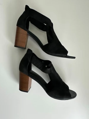 Vagabond skor i äkta läder - Ett par svarta högklackade skor / sandaler från Vagabond med öppen tå och träfärgade klackar. Äkta läder / skinn. Mycket gott skick, som nya.   Hämtas i Stockholm eller skickas mot porto  