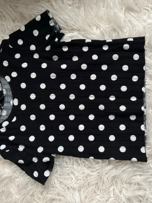 Svart prickig topp från Zara - Kortärmad topp från Zara i svart med vita prickar. Ribbad struktur och rund halsringning. Perfekt för dig som gillar ett lekfullt mönster och vill ha en enkel men snygg topp till jeans eller kjol.