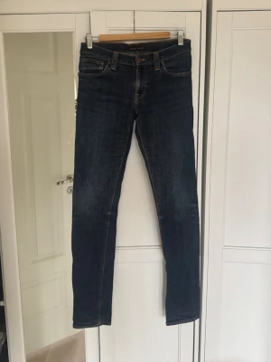 Mörkblå jeans från Nudie Jeans - Säljer ett par mörkblå jeans från Nudie Jeans med kontrastsömmar. Jeansen har raka ben och snygga broderade bakfickor med Nudie-loggan. STORLEK: W28, L34