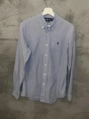 Ralph lauren skjorta - Klassisk blårutig skjorta från Ralph Lauren i modellen Custom Fit. Skjortan har button-down krage, vita knappar och den ikoniska broderade loggan på bröstet. Tillverkad i mjuk bomull och har lång ärm. Perfekt för en clean och stilren look.