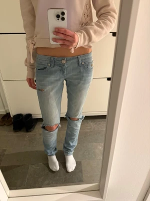 Ljusblå lågmidjade jeans med hål/slitningar/revor - Ljusblå jeans med låg midja. Jeansen har slitningar och hål vid knäna för en vintage look. Klassisk femficksmodell med snygga detaljer på fickorna och gul brodyr. Perfekta för dig som gillar en avslappnad stil.