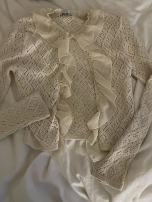 Beige kofta med volang från Zara - PRIS KAN DISKUTERAS!! Supersöt beige kofta från Zara med stickat hålmönster och stora volangdetaljer längs krage och framkant. Långärmad och i mjukt material som känns både romantiskt och trendigt. Perfekt att slänga över en topp för en extra touch. 