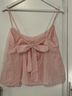 Ljusrosa linnetopp med rosett - Supersöt ljusrosa linnetopp med tunna axelband och stor rosett baktill. Toppen har en luftig och flowy passform, perfekt för varma dagar. Materialet är lätt och har en diskret struktur som ger en avslappnad vibe.