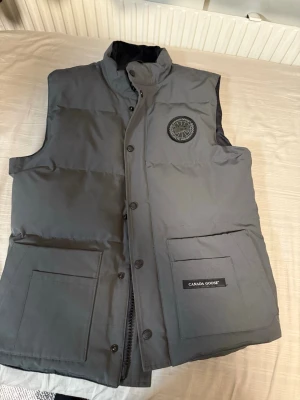 Grå dunväst från Canada Goose - Säljer en grå dunväst från Canada Goose med hög krage och stora fickor framtill. Västen har ett broderat svart märke på bröstet och logotyp på ena fickan. Perfekt för lager-på-lager och riktigt snygg till streetwear-stilen.                           Var fri att kasta ett bud de skadar ej är du intresserad.