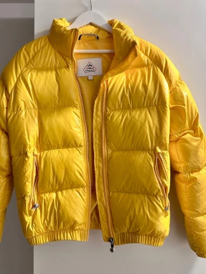 Gul pufferjacka från Pyrenex - Säljer en riktigt fet gul pufferjacka från Pyrenex. Jackan har hög krage, två dragkedjefickor och klassisk Pyrenex-logga på ärmen. Den är vadderad och har elastiska muddar vid ärmslut och midja. Perfekt för kalla dagar när du vill sticka ut.