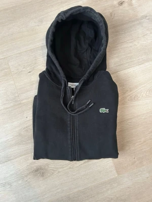 Svart Lacoste Zip Hoodie - Fin Lacoste hoodie i svart färg. Tröjan är i mycket bra skick. Sparsamt använd! |  Storlek: M | Skick: Mycket bra skick! | Färg: Svart | Märke: Lacoste | Modell: Hoodie med dragkedja |  Hör gärna av dig vid frågor så svarar jag så fort jag kan! 😁| 
