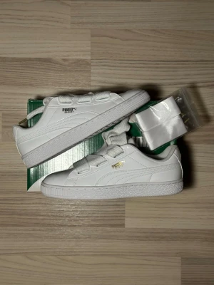 Vita Puma Basket sneakers med breda snören - Säljer ett par klassiska Puma Basket sneakers i helvitt skinn med breda, platta snören och extra satinsnören. Skorna har rund tå, platt sula och subtila gulddetaljer vid loggan. Perfekta för dig som gillar clean och stilren look.