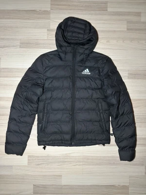 Svart pufferjacka från Adidas XS - Säljer en svart pufferjacka från Adidas i storlek XS. Jackan har huva, dragkedja framtill och två sidofickor. Adidas-logga på bröstet och justerbar nederkant. Perfekt för kalla dagar och riktigt skön att bära.