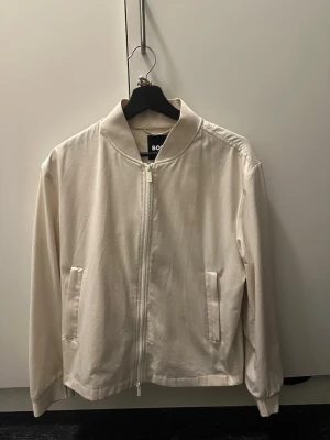 Hugo boss jacka - nyskick, använd 1 gång - Säljer en stilren och tidlös jacka från Hugo Boss i ljus/beige ton. Använd endast 1 gång, i princip som ny utan slitage eller fläckar. Väldigt snygg passform och lätt material – perfekt till vår/sommar eller som en tunn jacka inomhus. Säljs då den tyvärr inte kommer till användning, kvitto finns.