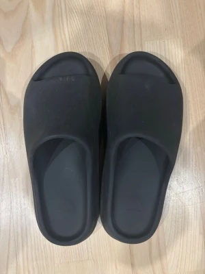 Svarta Yezzy slide - Säljer ett par svarta Adidas slides med chunky, räfflad sula och minimalistisk design. Tillverkade i syntetmaterial för en mjuk och bekväm känsla. Perfekta för chill dagar eller när du vill ha något enkelt att slänga på dig.