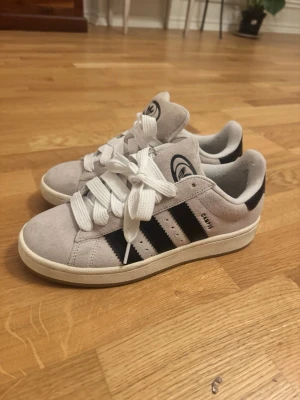 Adidas Campus sneakers i mocka - Säljer ett par Adidas Campus sneakers i ljusgrå mocka med klassiska svarta ränder på sidorna och vit sula. Skorna har rund tå, vita snören och Adidas-logga på plösen. Perfekta för dig som gillar streetstyle.