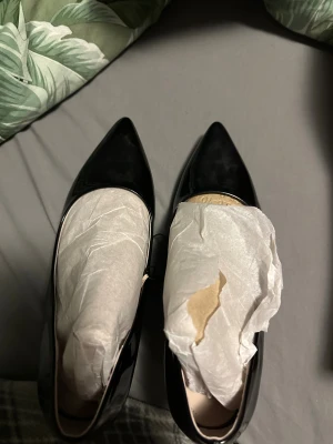 Svarta pumps med spetsig tå, storlek 39 - Säljer ett par svarta pumps i storlek 39 med glansig finish och spetsig tå. Skorna har hög klack och en elegant, smal siluett. Materialet är syntet med lackad yta som ger en snygg look. Perfekt för dig som gillar klassisk stil med edge.