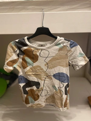 Mönstrad t-shirt från H&M, strl 122/128 - Snygg t-shirt från H&M i storlek 122/128 med abstrakt mönster i beige, blått, svart och brunt. T-shirten har rund halsringning och korta armar. Tillverkad i mjuk bomull, perfekt för en chill och trendig look. Barn till pojkar 