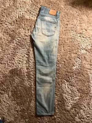 Replay Anbass ljusa jeans - Säljer ett par Replay Anbass jeans i ljusblå tvätt med snygga slitningar och klassiska fem fickor. Jeansen har röd detalj vid fickan, rak passform och normal midja. Perfekt för dig som gillar en avslappnad men stilren look.