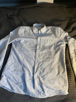 Randig Oxfordskjorta från ASKET XS - Snygg Oxfordskjorta från ASKET i storlek XS. Skjortan är vit med tunna blå ränder, har klassisk krage, bröstficka och knappar framtill. Tillverkad i mjuk bomull som känns skön mot huden. Perfekt för dig som gillar stilrena och fräscha plagg. Nypris 3000kr nästan och mitt pris 399!