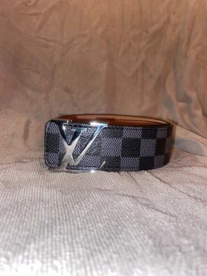 Louis Vuitton rutig skinnbälte - Säljer ett Louis Vuitton bälte med klassiskt rutigt mönster i svart och grått. Bältet har en stor silverfärgad LV-logga som spänne och är tillverkat i äkta skinn med ljusbrun insida. Perfekt accessoar för dig som gillar exklusiva detaljer.