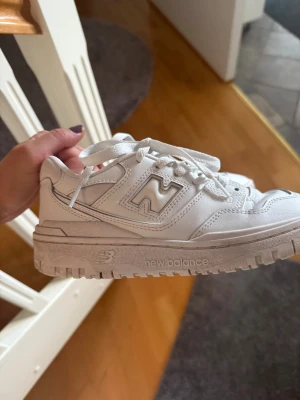 Vita New Balance sneakers - Säljer ett par fräscha vita New Balance sneakers med chunky platt sula och snörning. Skorna har en rund tå och är tillverkade i skinn och syntetmaterial. Klassisk New Balance-logga på sidan och detaljerade sömmar för en sportig vibe.