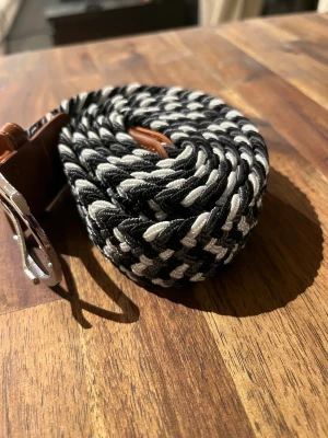 Svartvit randigt flätat bälte - Säljer ett nytt snyggt flätat bälte med svartvita ränder och detaljer i brunt läder. Bältet har en silverfärgad metallspänne och är gjort i ett slitstarkt syntetmaterial. Perfekt för att ge din outfit en cool touch med sitt mönstrade och trendiga utseende.