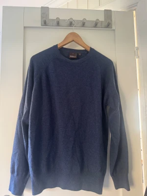 Mörkblå stickad tröja från Oscar jacobson - Snygg mörkblå stickad tröja från Oscar jacobson med rund hals och ribbade muddar vid ärmslut och nederkant. Perfekt för dig som gillar klassisk stil med en modern touch. Materialet är mjukt och värmande, passar dig som vill ha en enkel men stilren look. Tröjan är oanvänd och i nyskick.