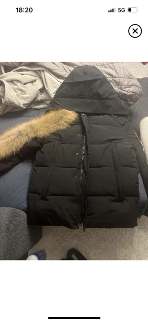 Svart pufferjacka med pälsdetalj - Säljer en svart pufferjacka med stor huva och snygg pälsdetalj längs kragen. Jackan har dragkedja och tryckknappar framtill samt två stora fickor. Perfekt för kalla dagar och har en stilren look som passar till det mesta.