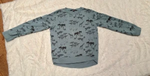 Blå sweatshirt med djurmönster från Lindex  - Säljer en blå sweatshirt från Lindex  med coolt djurmönster i svart, där giraffer, leoparder och noshörningar syns bland palmer och träd. Tröjan har långärmad passform, rund hals och ribbade muddar. Perfekt för dig som gillar safari vibes! Mycket bra skick förutom 2 ljusa fläckar på framsidan som syns tydligast på sista bild.