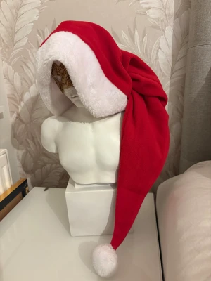 Röd tomteluva med vit kant och boll - Klassisk tomteluva i röd fleece med mjuk vit kant och fluffig vit boll längst ut. Luvan har en lång, spetsig form och är perfekt för julens alla tillfällen. Materialet är mjukt och skönt mot huvudet, och den vita kanten ger en extra mysig känsla.