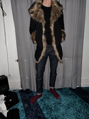 Svart parkajacka med fuskpäls - Säljer en svart parkajacka med stor huva och detaljer i brun fuskpäls längs krage, ärmslut och öppning. Jackan har en längre passform och är perfekt för dig som vill sticka ut med en cool look. Materialet är syntet med mjuk pälsimitation.