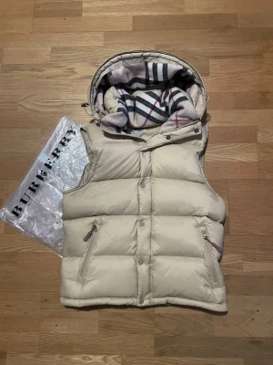 Beige dunväst från Burberry - Stilren beige dunväst från Burberry med avtagbara ärmar och klassiskt rutigt foder i huvan. Västen har dragkedja och knappar framtill samt två fickor med dragkedja. Perfekt för lager-på-lager och ikonisk Burberry-look. Pris går att diskuteras.