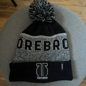 Svart och grå Örebro hockey mössa med tofs - Snygg svart och grå mössa med stor tofs på toppen och bred uppvikt kant. Merch från Örebro hockey.