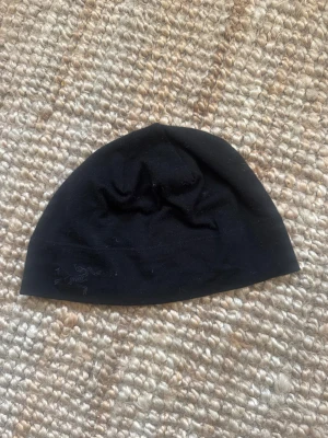 Svart mössa från Arc'teryx - Svart, stilren mössa från Arc'teryx med diskret broderad logga framtill. Mjuk och tunn design som sitter skönt på huvudet, perfekt för kyliga dagar. Materialet är merinoull som håller värmen utan att bli för tjockt.