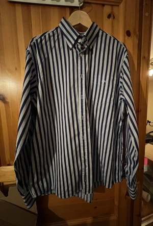 Randig skjorta från U.S. Polo Assn XL - Snygg skjorta från U.S. Polo Assn i storlek XL med breda marinblå och vita ränder. Klassisk button-down krage, bröstficka med broderad logga och knäppning framtill. Tillverkad i 100% bomull.