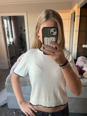Vit stickad topp med svarta detaljer - Säljer en vit stickad topp med svarta kanter vid halsringning, ärmslut och nederkant. Modellen är croppad och har korta ärmar. Perfekt för dig som gillar enkel men snygg stil. Materialet är mjukt och ger en chill vibe.