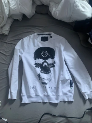 Vit Philipp Plein sweatshirt med döskalle - Säljer en vit sweatshirt från Philipp Plein i storlek L med ett stort svart döskalletryck och logga framtill. Tröjan har rund hals, långa ärmar och ribbade muddar. Materialet är mjuk bomull och designen är riktigt edgy med coola detaljer.