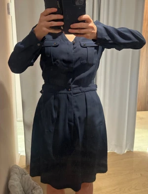 Marinblå klänning Bondelid XS - Snygg och skön marinblå klänning med v-ringning, långa ärmar och knappar framtill. Klänningen har två bröstfickor med lock och ett matchande bälte i midjan som ger en cool siluett. Materialet känns mjukt och har ett lätt fall. Perfekt för dig som gillar stilrena och klassiska plagg.