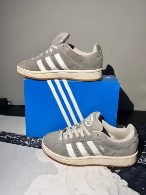 Adidas Campus 00s grå sneakers strl 38 - Adidas Campus 00s sneakers i grå mocka med vita klassiska stripes och gummisula. Skorna har rund tå, snörning och retro vibe. Adidas-logga på plösen och Campus-tryck på sidan. Perfekt för dig som gillar streetstyle och vill ha en clean look.