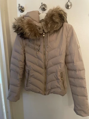 Beige pufferjacka med fuskpäls från Hollies - Snygg beige pufferjacka från Hollies med fluffig fuskpäls på huvan och leopardmönstrat innerfoder. Jackan har flera dragkedjor i guld, snörning vid huvan och quiltad design. Perfekt för dig som vill ha både stil och värme under vintern.